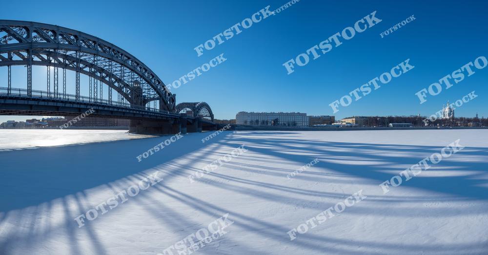 Фотография зимнего городского пейзажа в Санкт-Петербург, Россия. На изображении запечатлён Большеохтинский мост, также известный как мост Петра Великого, на фоне голубого неба. Фотоснимок передаёт атмосферу заснеженного Петербурга: мощные металлические конструкции моста отбрасывают выразительную тень, подчёркивая инженерную архитектуру и масштаб сооружения. Фото отражает сочетание зимней природы и индустриального облика города, а также вид на реку Нева. Иллюстрация может использоваться как картинка или слайд, посвящённые городам России и архитектуре мостов Санкт-Петербурга.